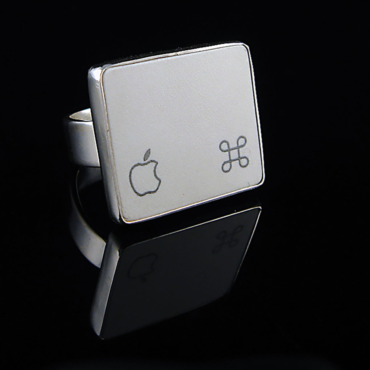 A silver ring featuring a white square with white Apple logo and command symbol, photographed on a clean glossy black background.