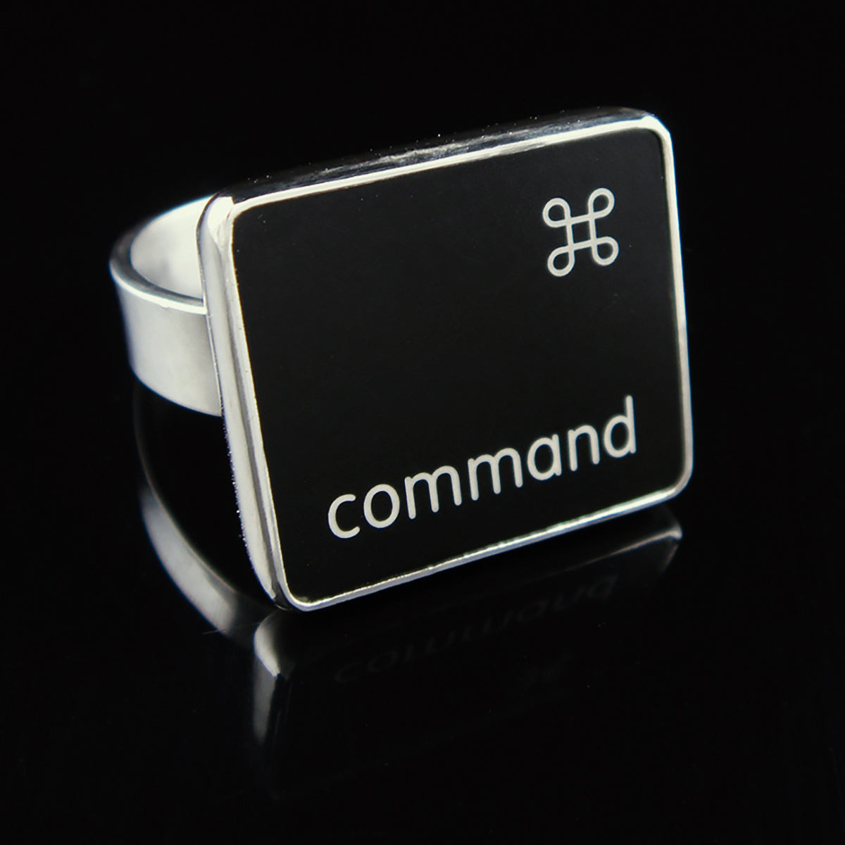 A silver ring featuring a black square with the word 'command' and the command small symbol, photographed on a glossy black reflective surface.