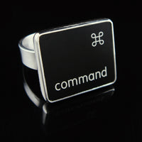 A silver ring featuring a black square with the word 'command' and the command small symbol, photographed on a glossy black reflective surface.