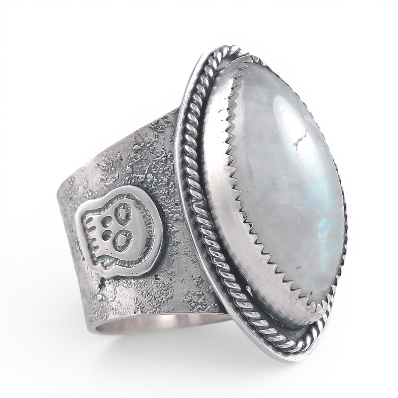 A wide silver ring featuring a large oval diamond cabochon moonstone with a soft gray-blue iridescent sheen, set in a textured band with a decorative skull motif and rope-style border.