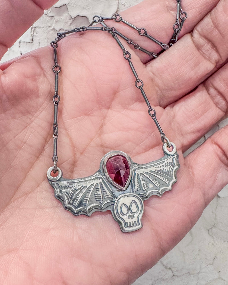 Hand holding a handmade pendant with a skull and bat wings and a teardrop shaped garnet. 