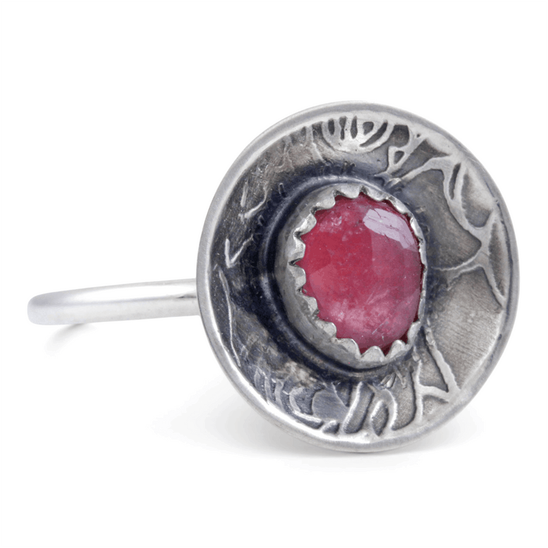 A sterling silver ring featuring a round, rose pattern textured dome set with a faceted pinkish-red gemstone, shown on a plain white background.