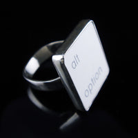 A silver ring featuring a white vertical rectangle with the lowercase letters 'alt' and 'option', photographed on a reflective black surface.