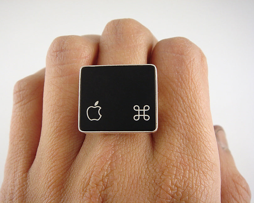 A close-up of a hand wearing a square black ring with a silver-tone border, featuring the Apple logo on one side and a command symbol on the other.