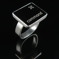 A silver ring featuring a black square with the word 'command' and the command small symbol, photographed on a glossy black reflective surface.