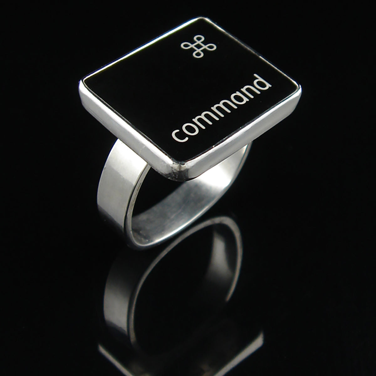A silver ring featuring a black square with the word 'command' and the command small symbol, photographed on a glossy black reflective surface.