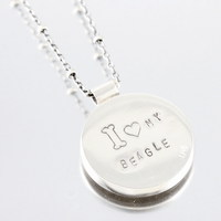 Back of a silver round pendant necklace engraved with 'I <heart symbol> MY BEAGLE', shown on a light gray reflective background.