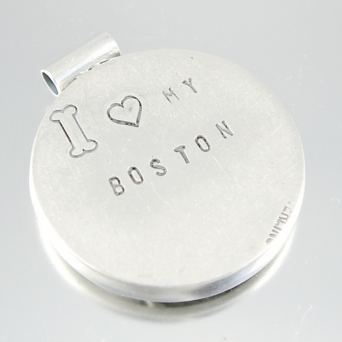 A round silver-toned metal keepsake or locket with a small bail at the top, engraved with a bone, a heart, and the words 'I ♥ MY BOSTON'.