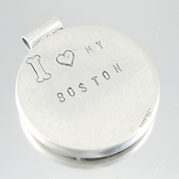 A round silver-toned metal keepsake or locket with a small bail at the top, engraved with a bone, a heart, and the words 'I ♥ MY BOSTON'.
