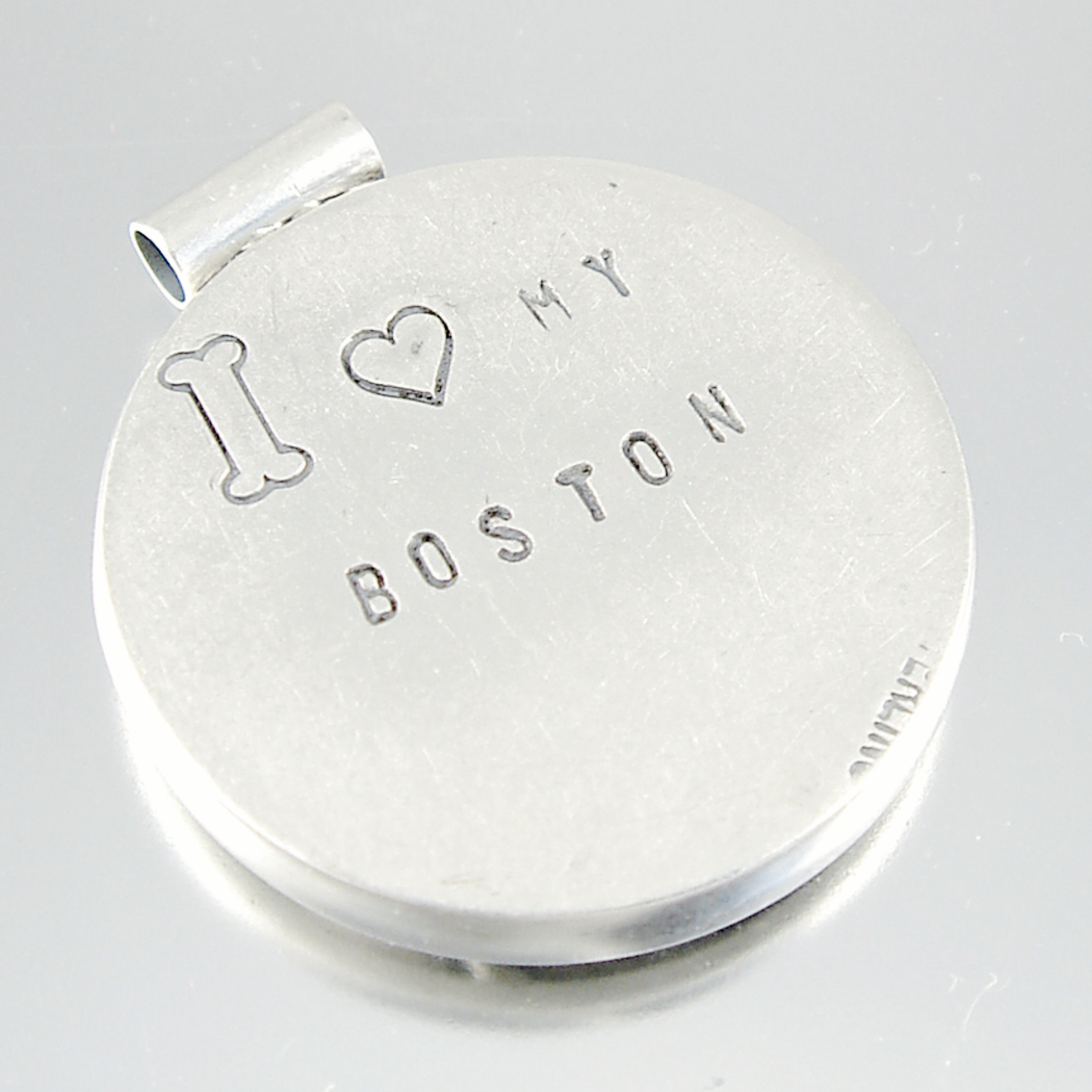 A round silver-toned metal keepsake or locket with a small bail at the top, engraved with a bone, a heart, and the words 'I ♥ MY BOSTON'.