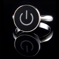 A silver ring featuring a round black face with a white on/off symbol, displayed on a reflective black surface.