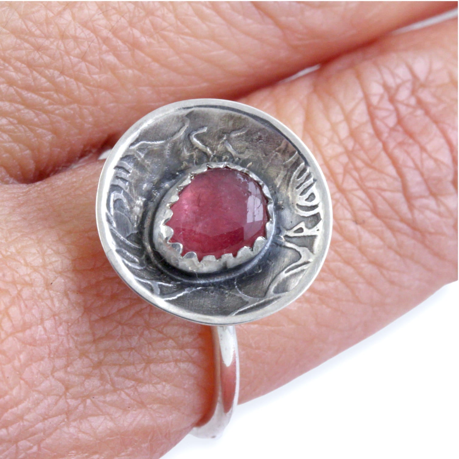 A sterling silver ring featuring a round, rose pattern textured dome set with a faceted pinkish-red gemstone, shown on a finger.