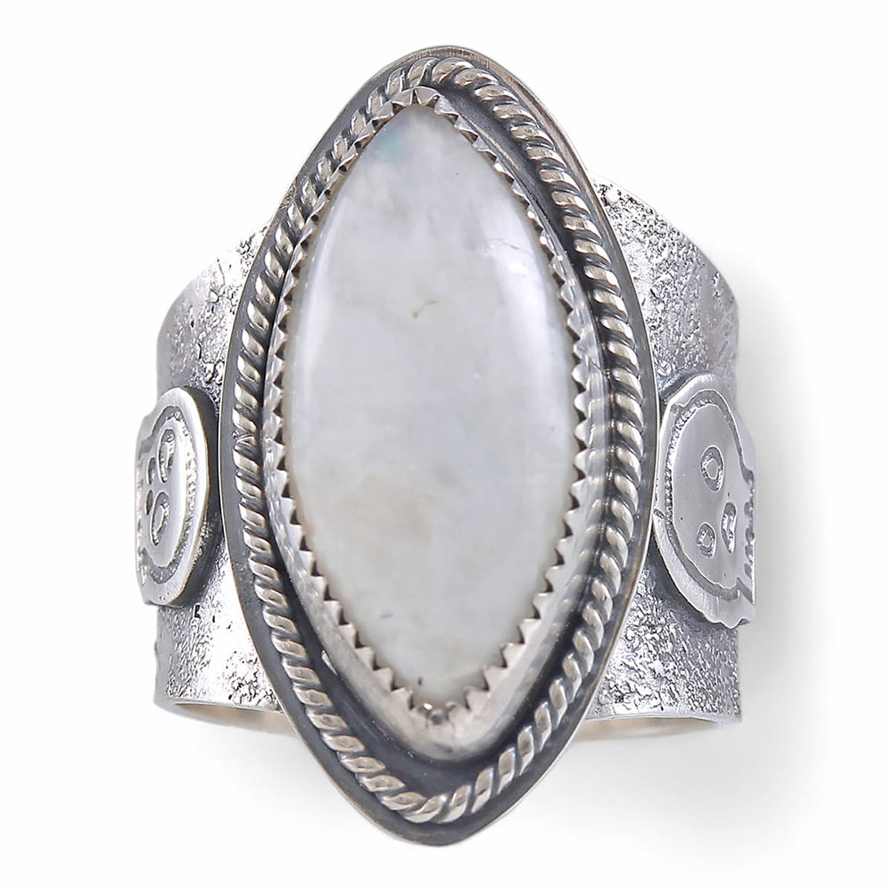 A wide silver ring featuring a large oval diamond cabochon moonstone with a soft gray-blue iridescent sheen, set in a textured band with a decorative skull motif and rope-style border.