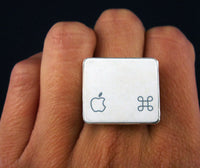 A silver square ring worn on a finger, featuring the Apple logo on one side and a geometric command symbol on the other, set against a dark background.