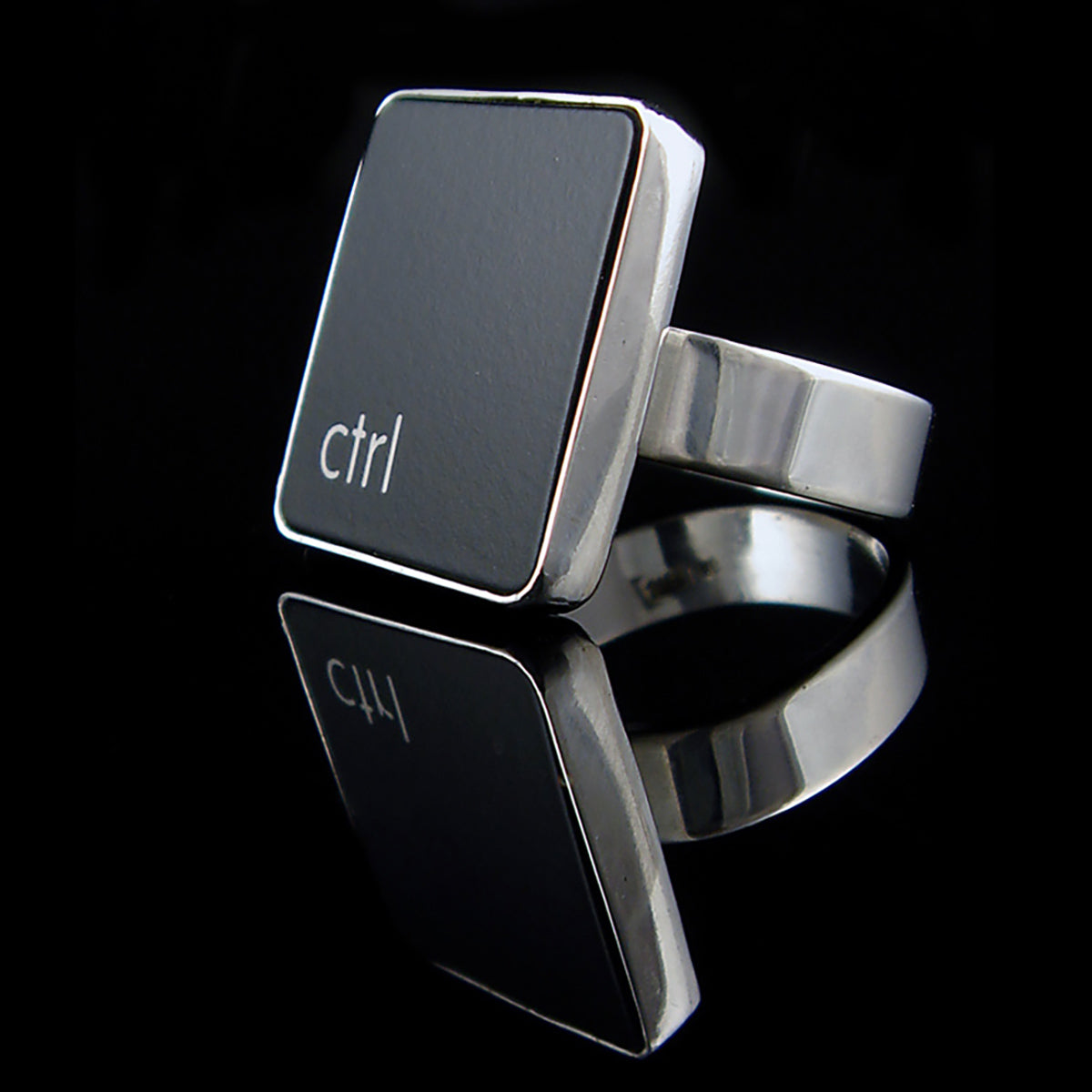 A silver ring featuring a black vertical rectangle with the lowercase letters 'ctrl', photographed on a reflective black surface.