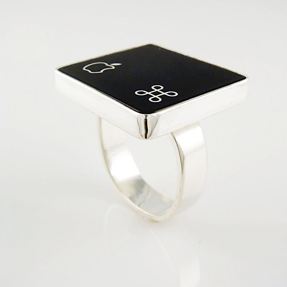 A silver ring featuring a black square with white Apple logo and command symbol, photographed on a clean white background.