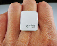 A close-up of a hand wearing a silver ring featuring a white vertical rectangle with the words 'enter' printed in white lowercase text.