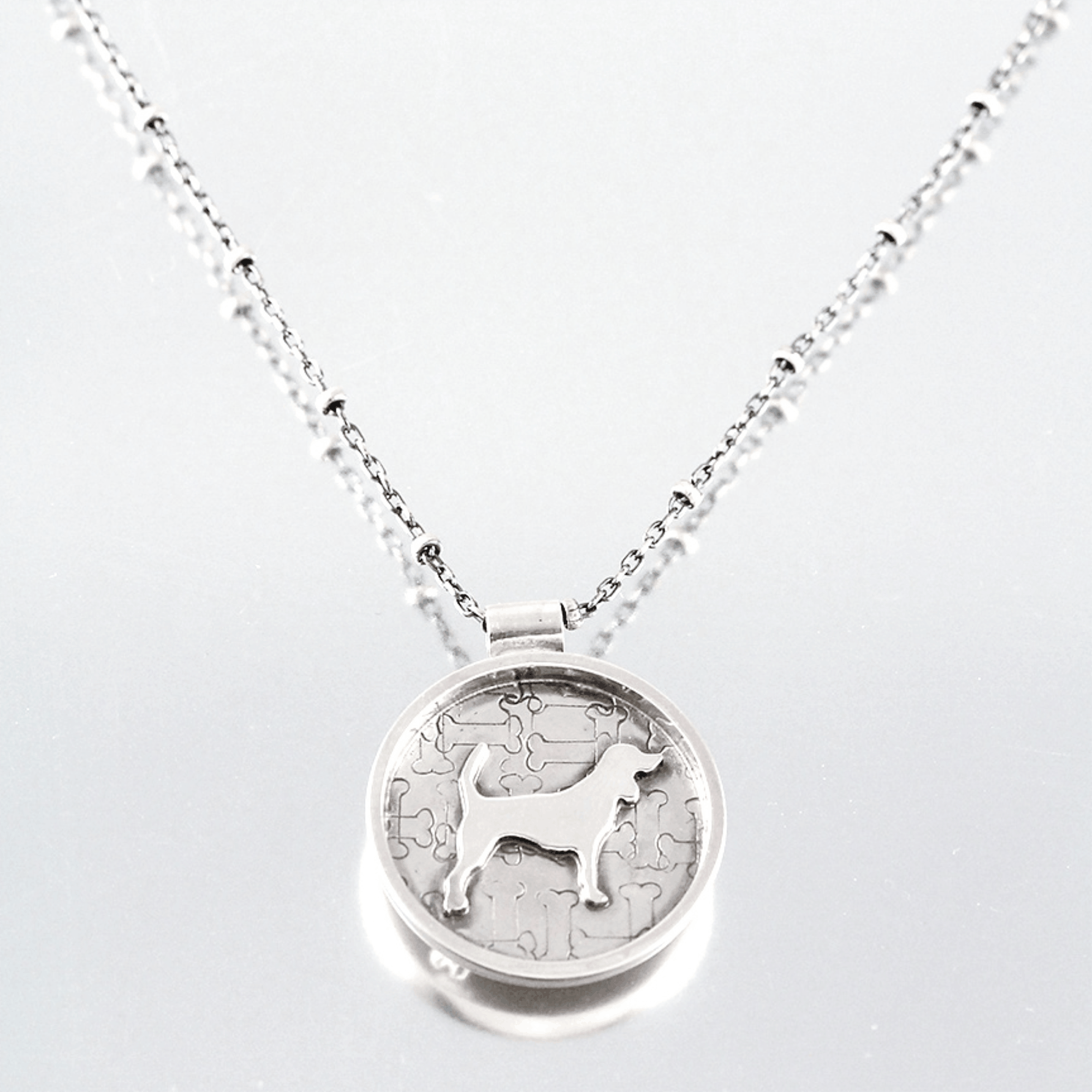 A silver round pendant necklace featuring a beagle dog silhouette over a jigsaw bone pattern, suspended from a silver chain on a reflective grey background.