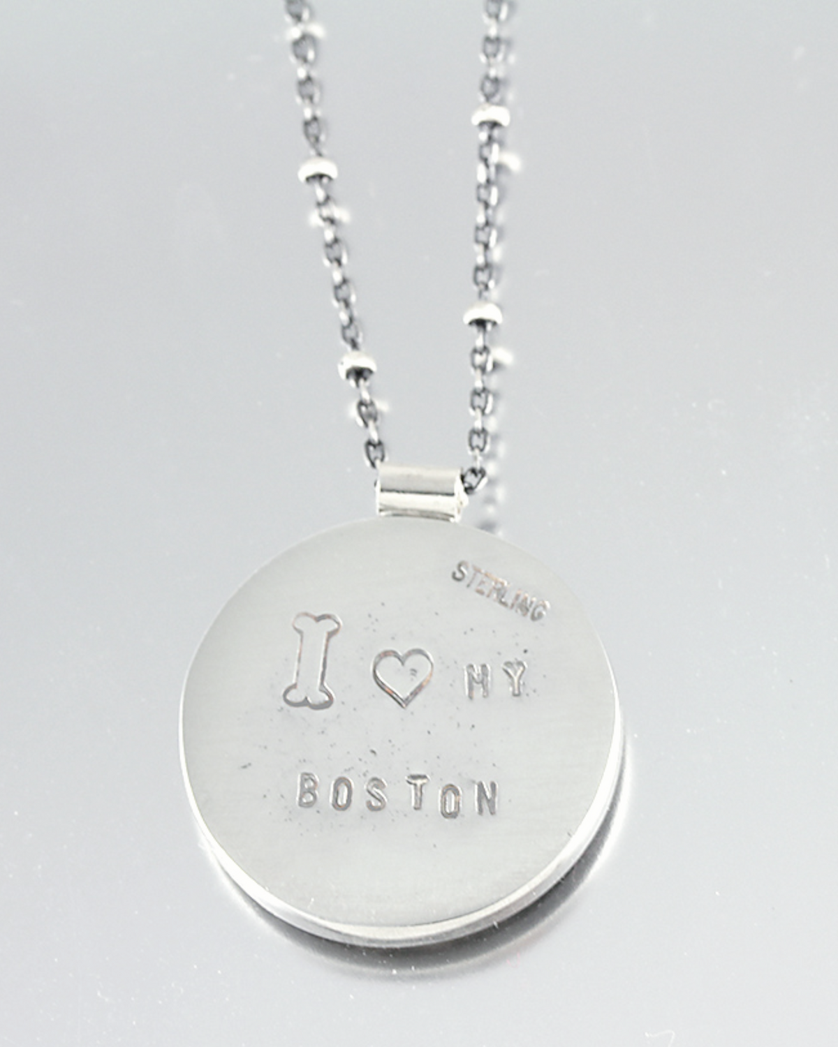 A sterling silver round pendant necklace engraved with 'I Heart MY BOSTON' and 'STERLING', shown on a light gray reflective surface.