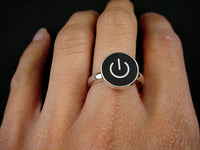 A silver ring with a black circular face featuring a white on/off symbol, shown worn on a person's finger against a dark background.