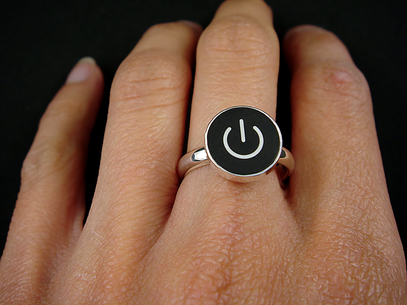 A silver ring with a black circular face featuring a white on/off symbol, shown worn on a person's finger against a dark background.