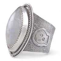 A wide silver ring featuring a large oval diamond cabochon moonstone with a soft gray-blue iridescent sheen, set in a textured band with a decorative skull motif and rope-style border.