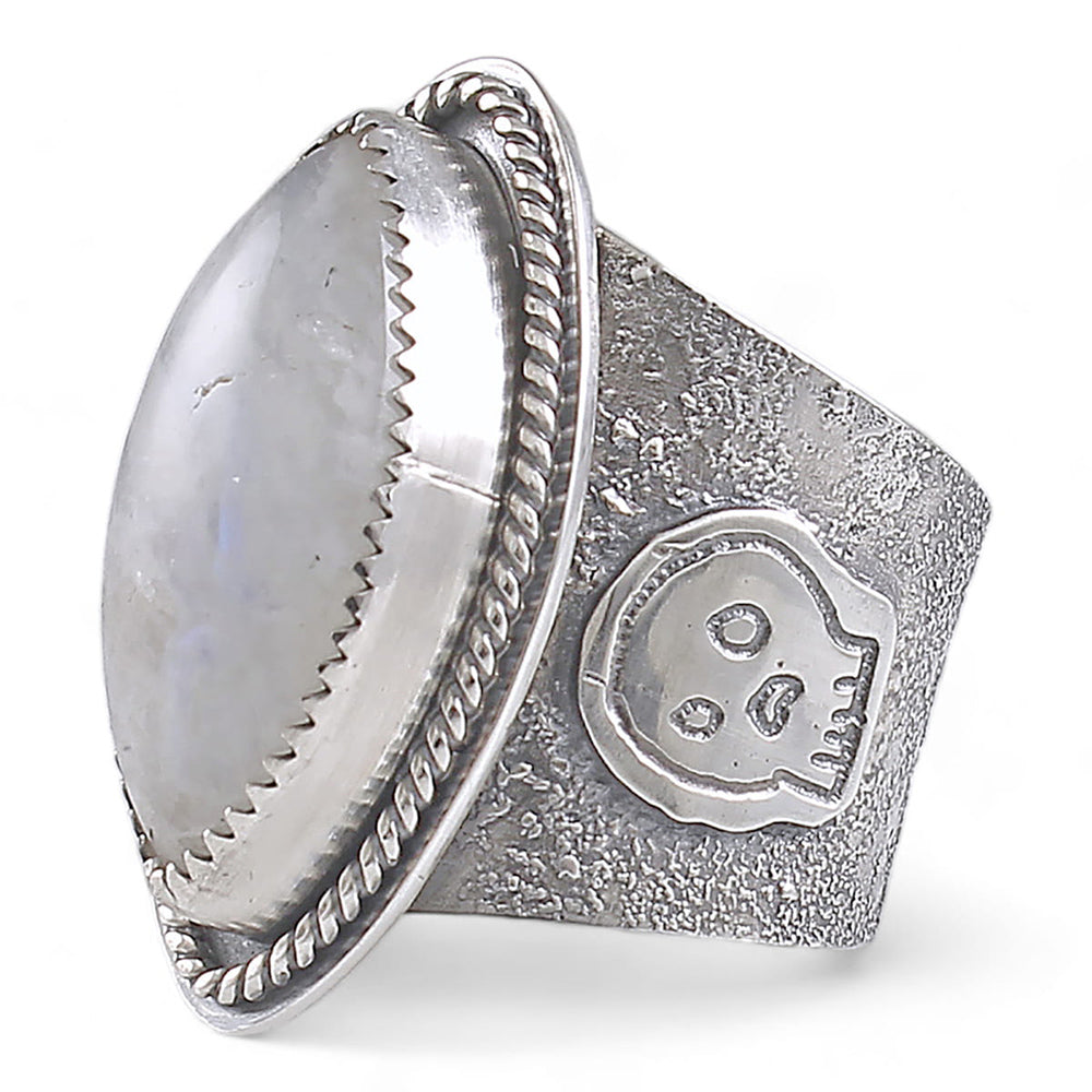 A wide silver ring featuring a large oval diamond cabochon moonstone with a soft gray-blue iridescent sheen, set in a textured band with a decorative skull motif and rope-style border.