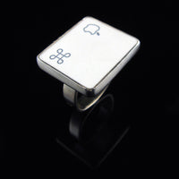 A silver ring featuring a white square with white Apple logo and command symbol, photographed on a clean glossy black background.