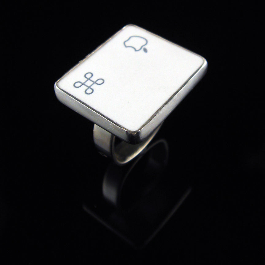 A silver ring featuring a white square with white Apple logo and command symbol, photographed on a clean glossy black background.
