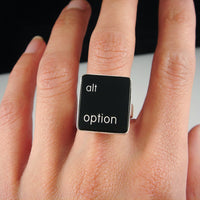 A close-up of a hand wearing a silver ring featuring a black vertical rectangle with the words 'alt option' printed in white lowercase text.