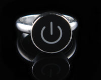 A silver ring featuring a round black face with a white on/off symbol, displayed on a reflective black surface.