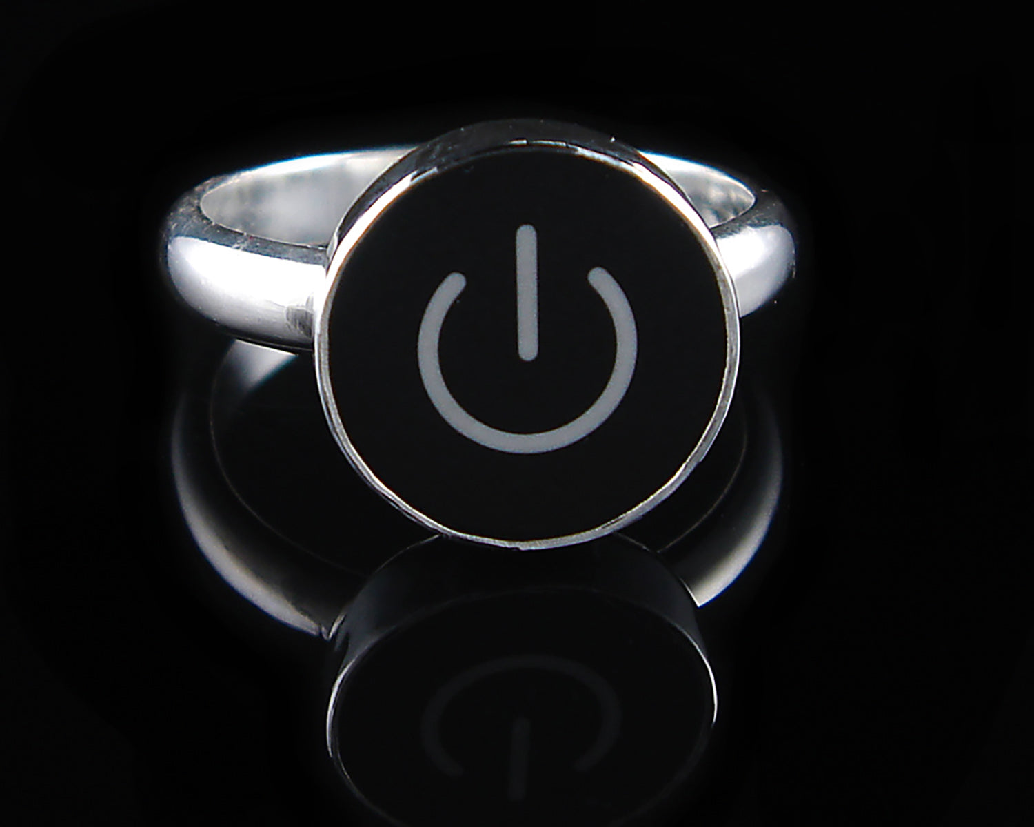 A silver ring featuring a round black face with a white on/off symbol, displayed on a reflective black surface.