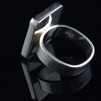 A polished sterling silver ring with a thick, rounded band and a large, flat, square-like side projection, displayed on a reflective black surface.