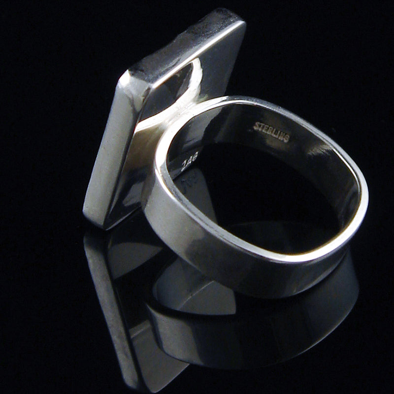 A polished sterling silver ring with a thick, rounded band and a large, flat, square-like side projection, displayed on a reflective black surface.