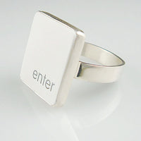 A silver ring featuring a white vertical rectangle with the lowercase letters 'enter', shown on a clean white reflective surface.