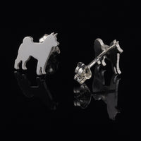 A pair of Shiba Inu silver dog silhouette stud earrings shown on a glossy black reflective surface, with one earring facing forward and the other angled to show the post and backing. 