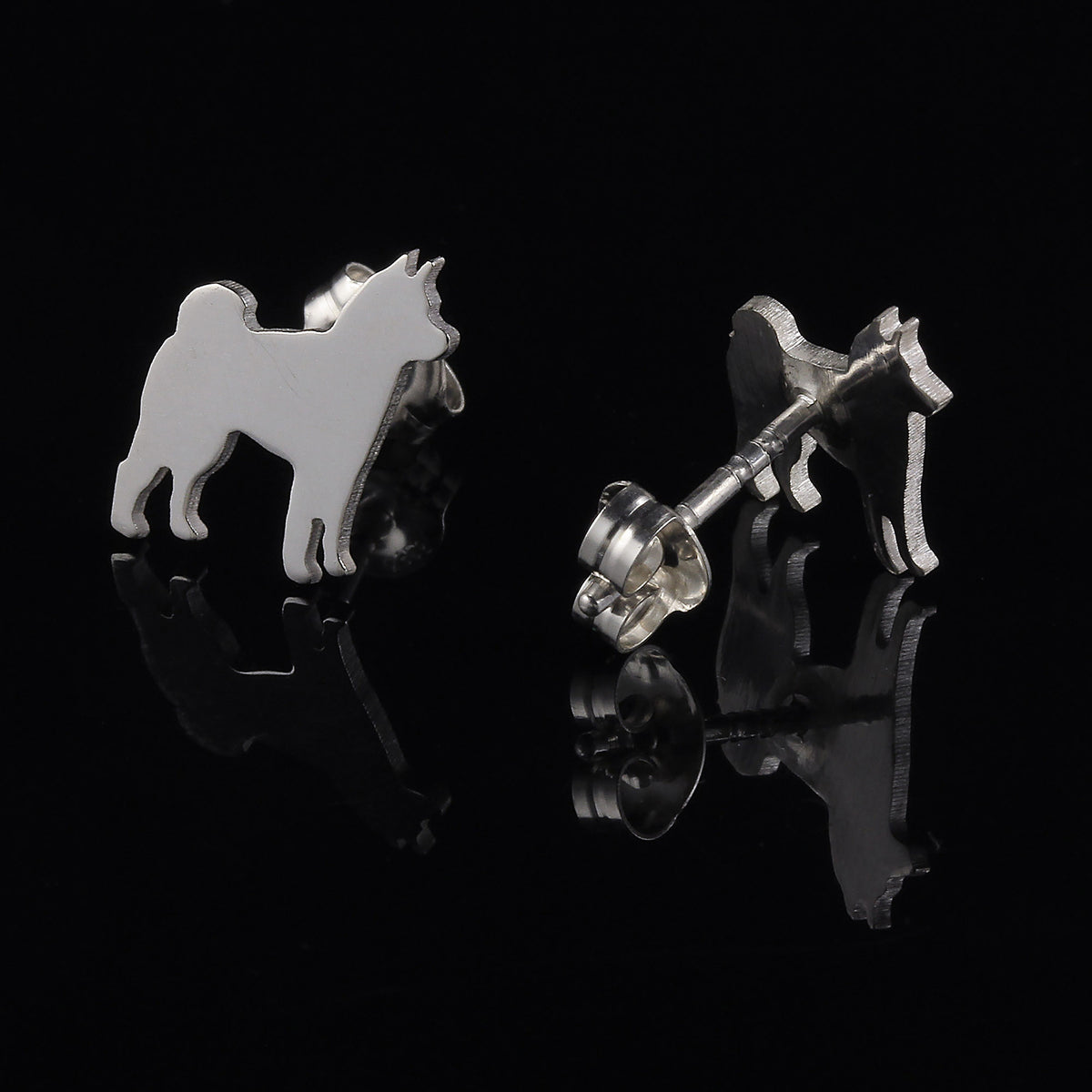 A pair of Shiba Inu silver dog silhouette stud earrings shown on a glossy black reflective surface, with one earring facing forward and the other angled to show the post and backing. 