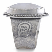 A wide silver ring featuring a large oval diamond cabochon moonstone with a soft gray-blue iridescent sheen, set in a textured band with a decorative skull motif and rope-style border.