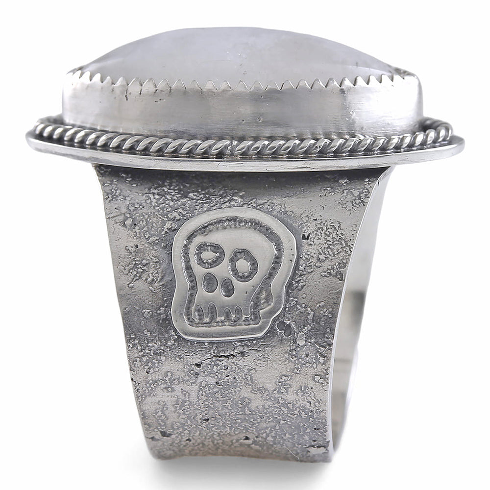 A wide silver ring featuring a large oval diamond cabochon moonstone with a soft gray-blue iridescent sheen, set in a textured band with a decorative skull motif and rope-style border.