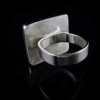 A silver ring with a wide, polished band and a square, textured face, displayed on a reflective black surface.