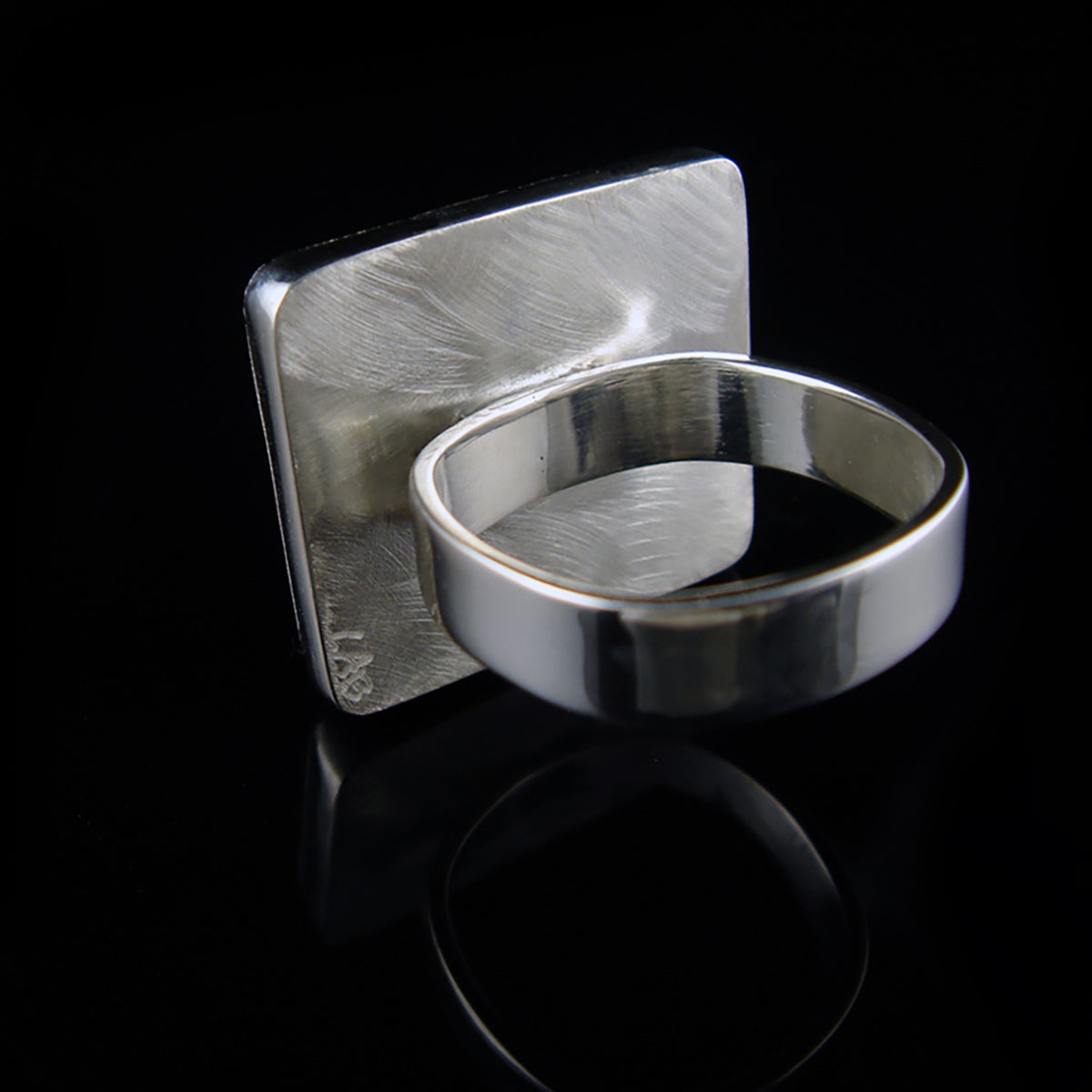 A silver ring with a wide, polished band and a square, textured face, displayed on a reflective black surface.
