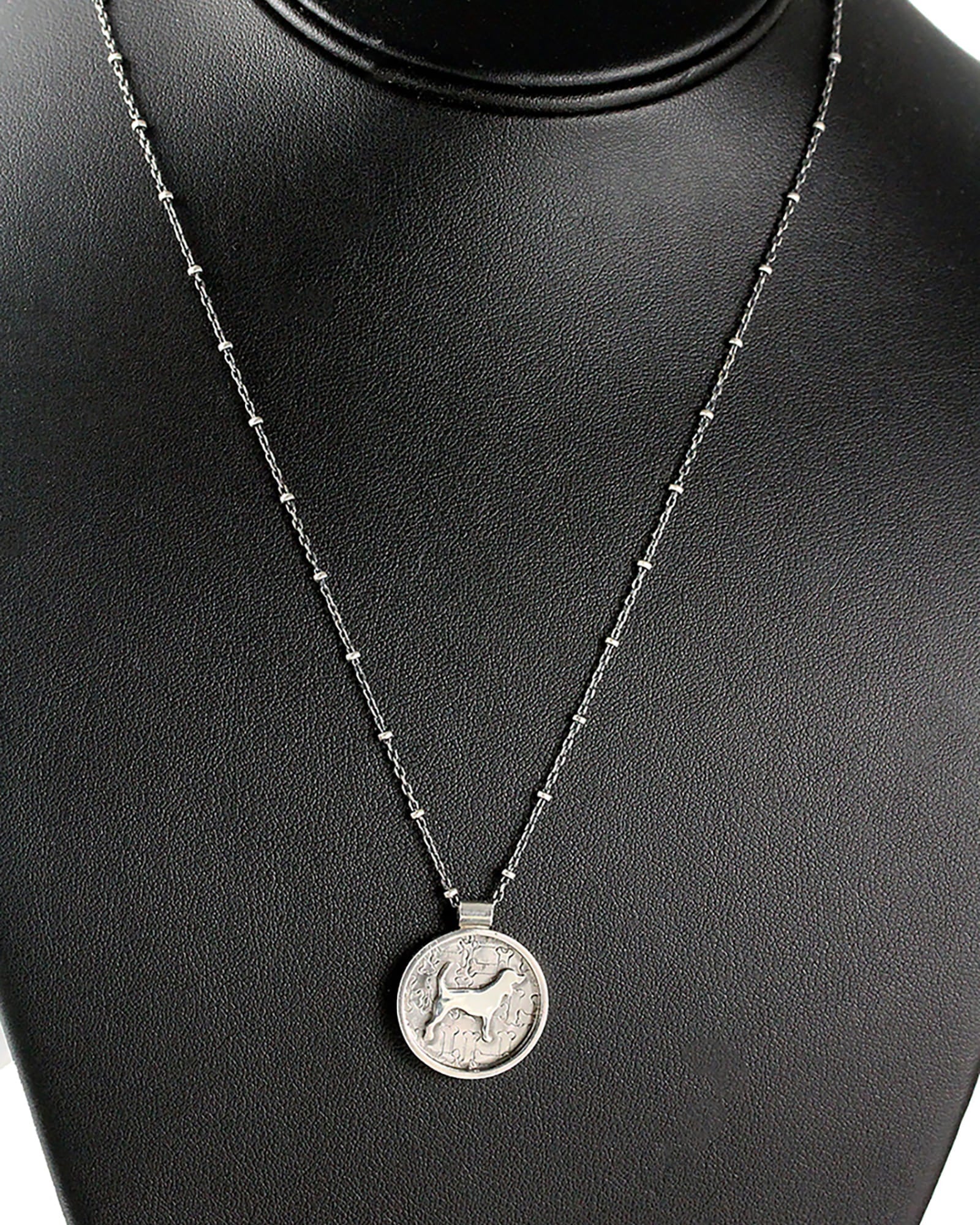 A silver coin pendant necklace featuring a beagle dog motif, displayed on a black textured jewelry bust.
