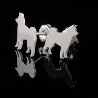 A pair of silver stud earrings shaped like Shiba Inu dog silhouettes, shown on a reflective black surface against a black background.