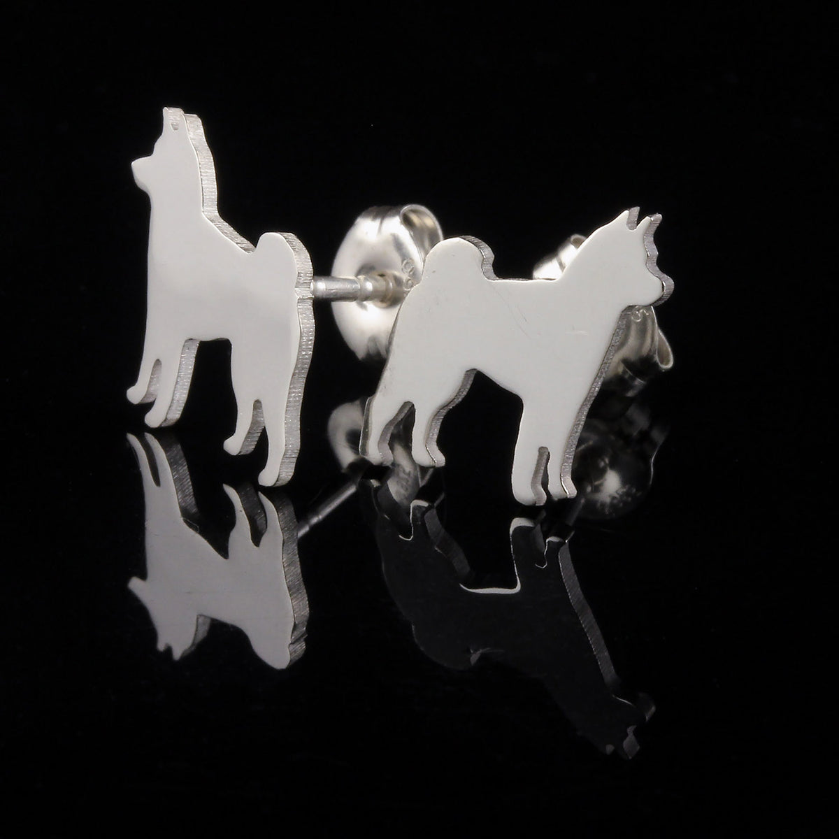 A pair of silver stud earrings shaped like Shiba Inu dog silhouettes, shown on a reflective black surface against a black background.