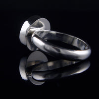 A polished silver metal ring with a wide, circular flat top, photographed on a reflective black surface.