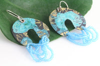 Colorful arch shaped dangle earrings with matte light blue beads on a natural green leaf on a white background.