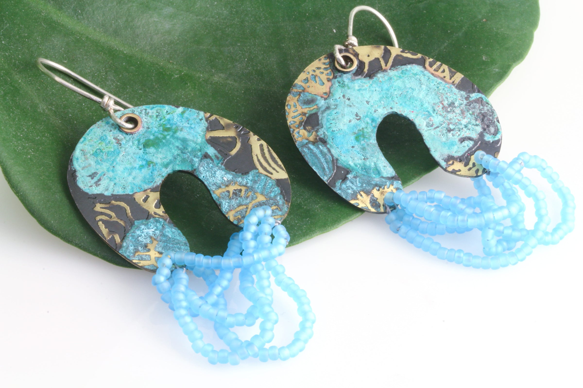 Colorful arch shaped dangle earrings with matte light blue beads on a natural green leaf on a white background.