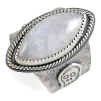 A wide silver ring featuring a large oval diamond cabochon moonstone with a soft gray-blue iridescent sheen, set in a textured band with a decorative skull motif and rope-style border.