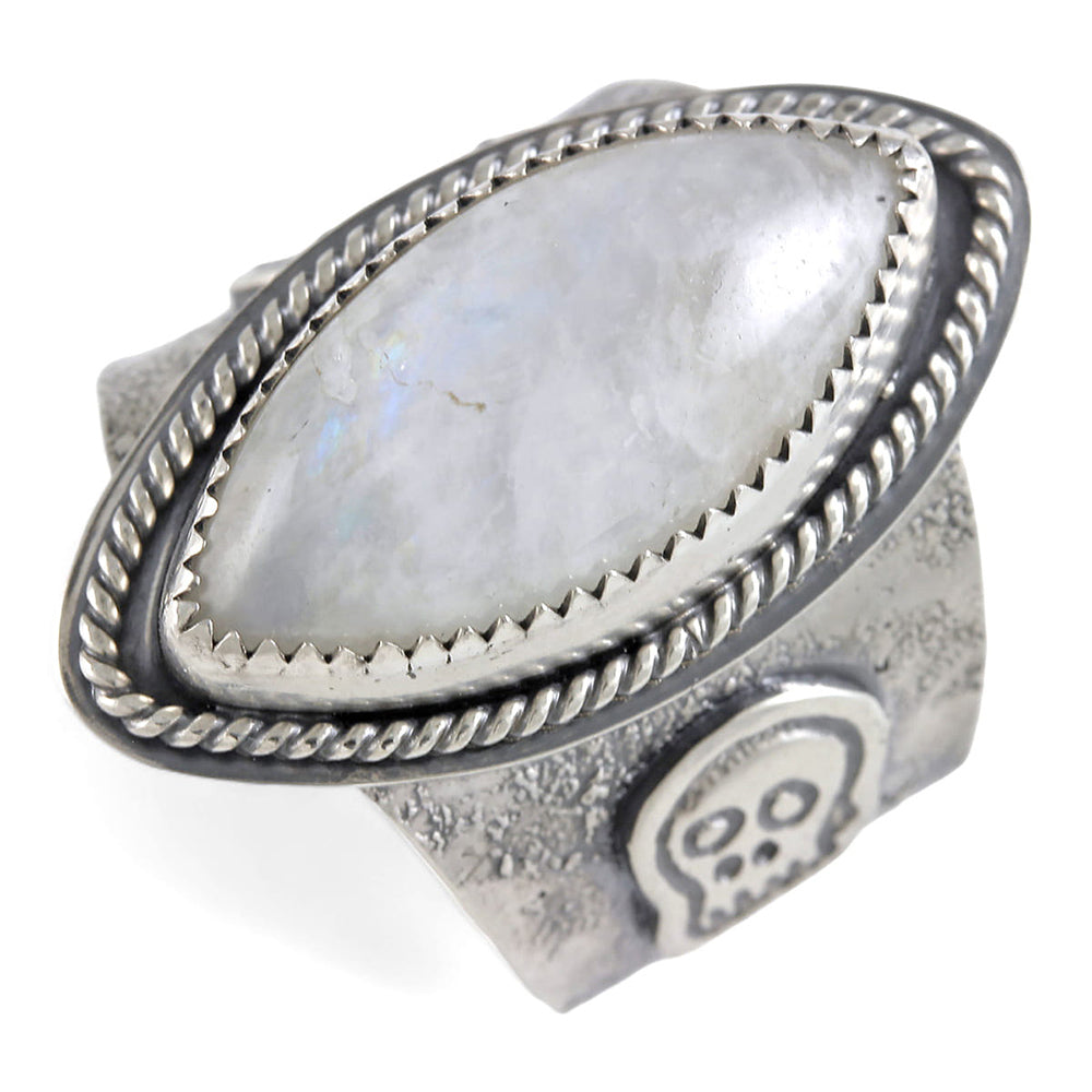 A wide silver ring featuring a large oval diamond cabochon moonstone with a soft gray-blue iridescent sheen, set in a textured band with a decorative skull motif and rope-style border.