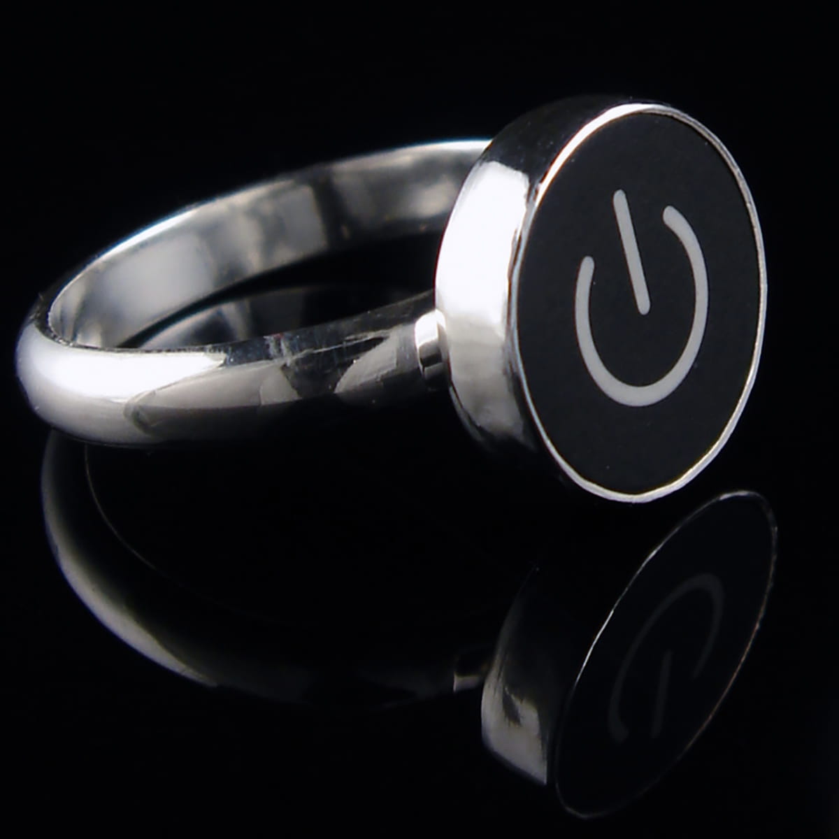 A silver ring featuring a round black face with a white on/off symbol, displayed on a reflective black surface.